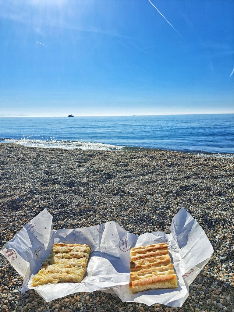 I am Sofia Lena Perissinotto, I am a blogger, content creator. In this article, I write about Celle Ligure, one of the most beautiful villages in Italy. This photo features a stunning view of the Mediterranean sea with a beach in Celle Ligure. It's early in the morning and I am having fresh focaccia for breakfast!