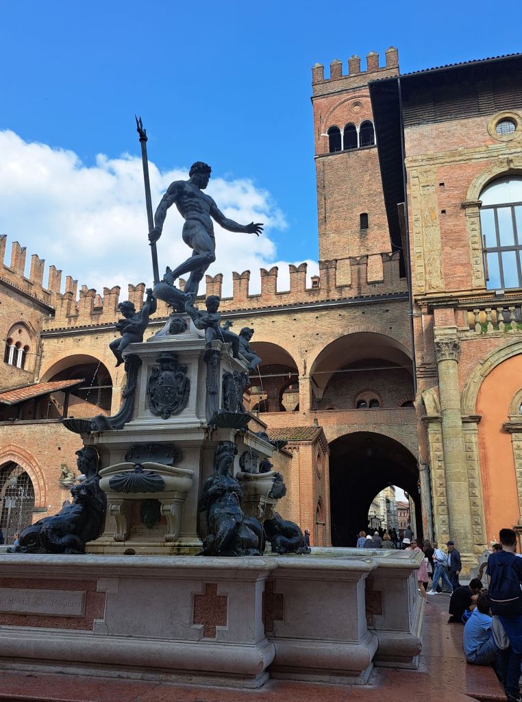 I'm Sofia Lena Perissinotto. I am a travel blogger. In this blog post I talk about Bologna, a stunning, historical city located in the Italian region Emilia-Romagna. The photo is taken in the stunning and most famous piazza of Bologna: Piazza Maggiore. The photo shows some buildings and the stunning marble fountain of Neptune.